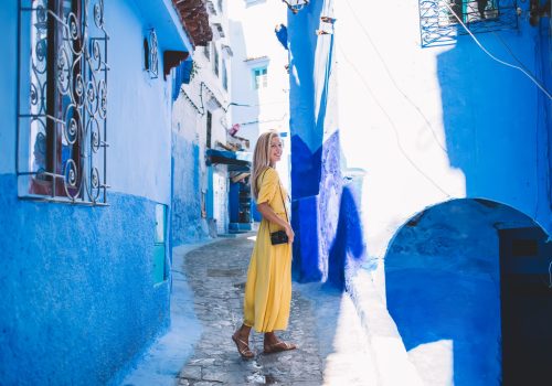 Full body glad female photographer smiling and looking at camera while standing on stone path outside shabby houses on sunny day in Morocco